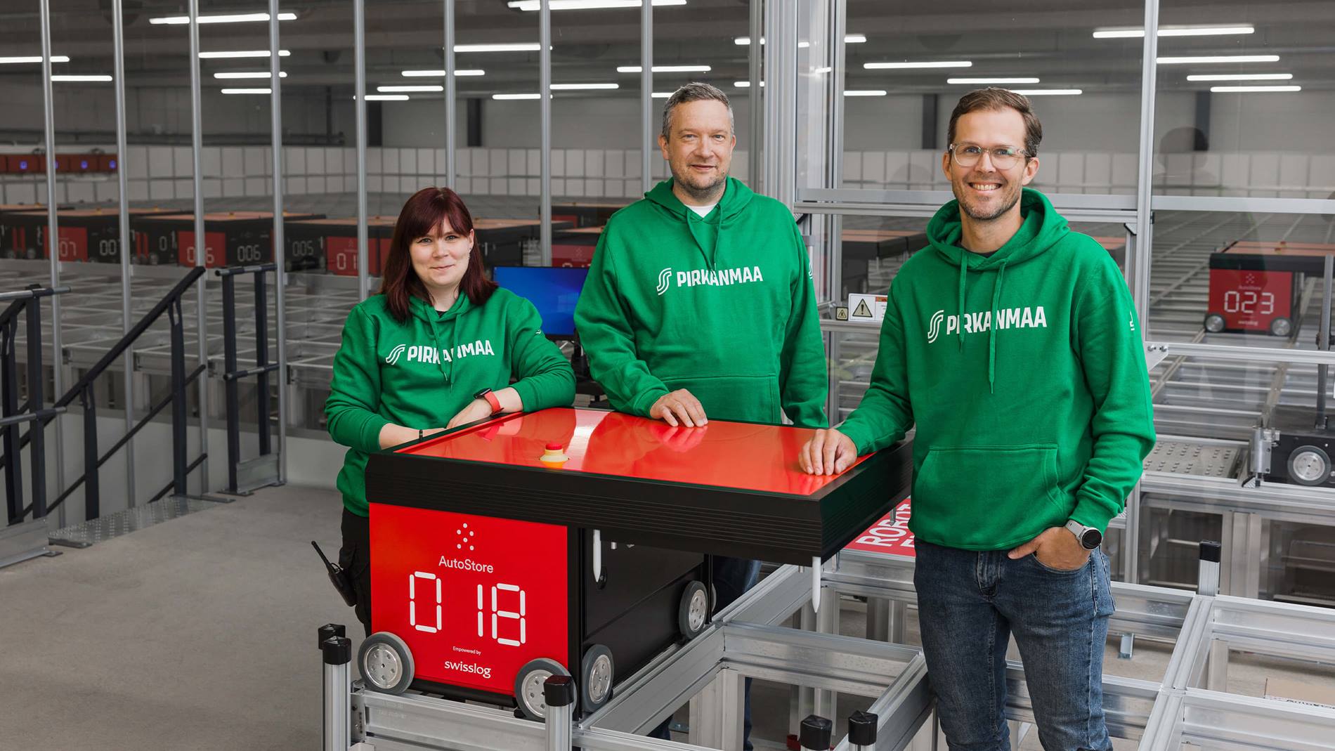 Kolme vihreään Pirkanmaan huppariin pukeutunutta henkilöä seisoo punaisen automaattisen varastointijärjestelmän robotin vieressä varastossa.
