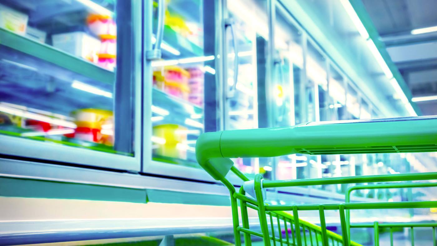 A shopping cart located in front of a refrigerator displaying frozen food products in a grocery store aisle.