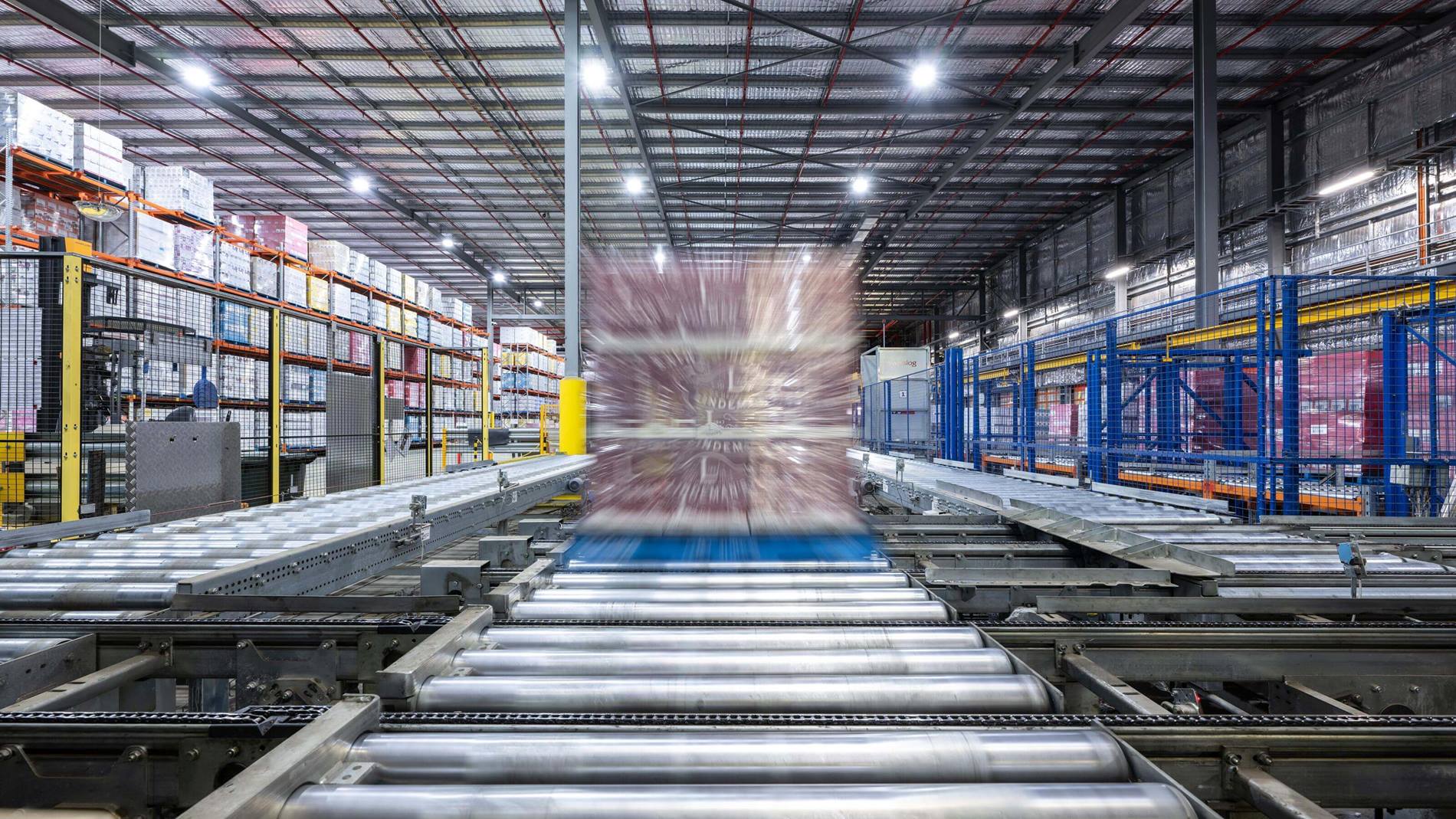 High-speed pallet conveyor system transporting a wrapped load through a modern warehouse with safety barriers and stocked shelves