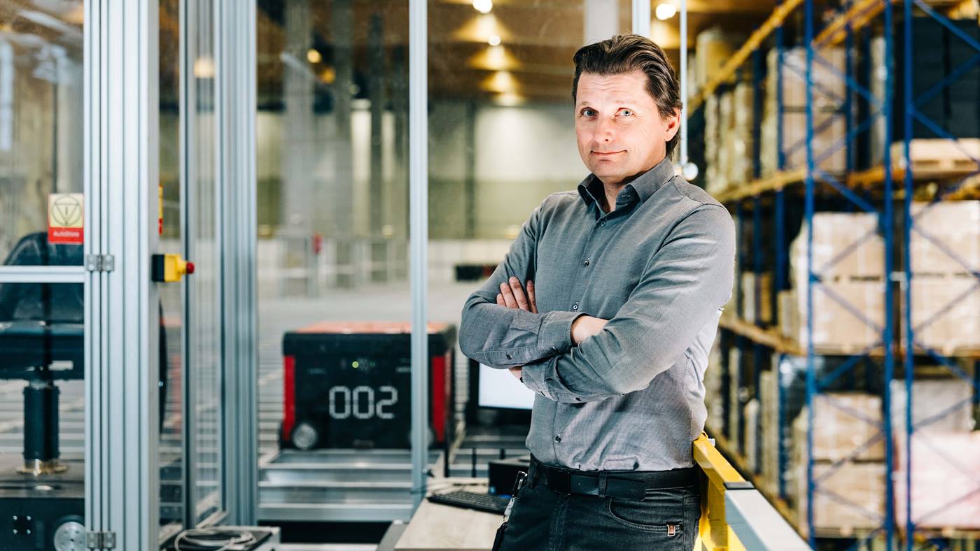 Tomas Skott from Nordic Lights, leaning against a railing with arms crossed. In the background, there are shelves with boxes, and an AutoStore robot on a grid.