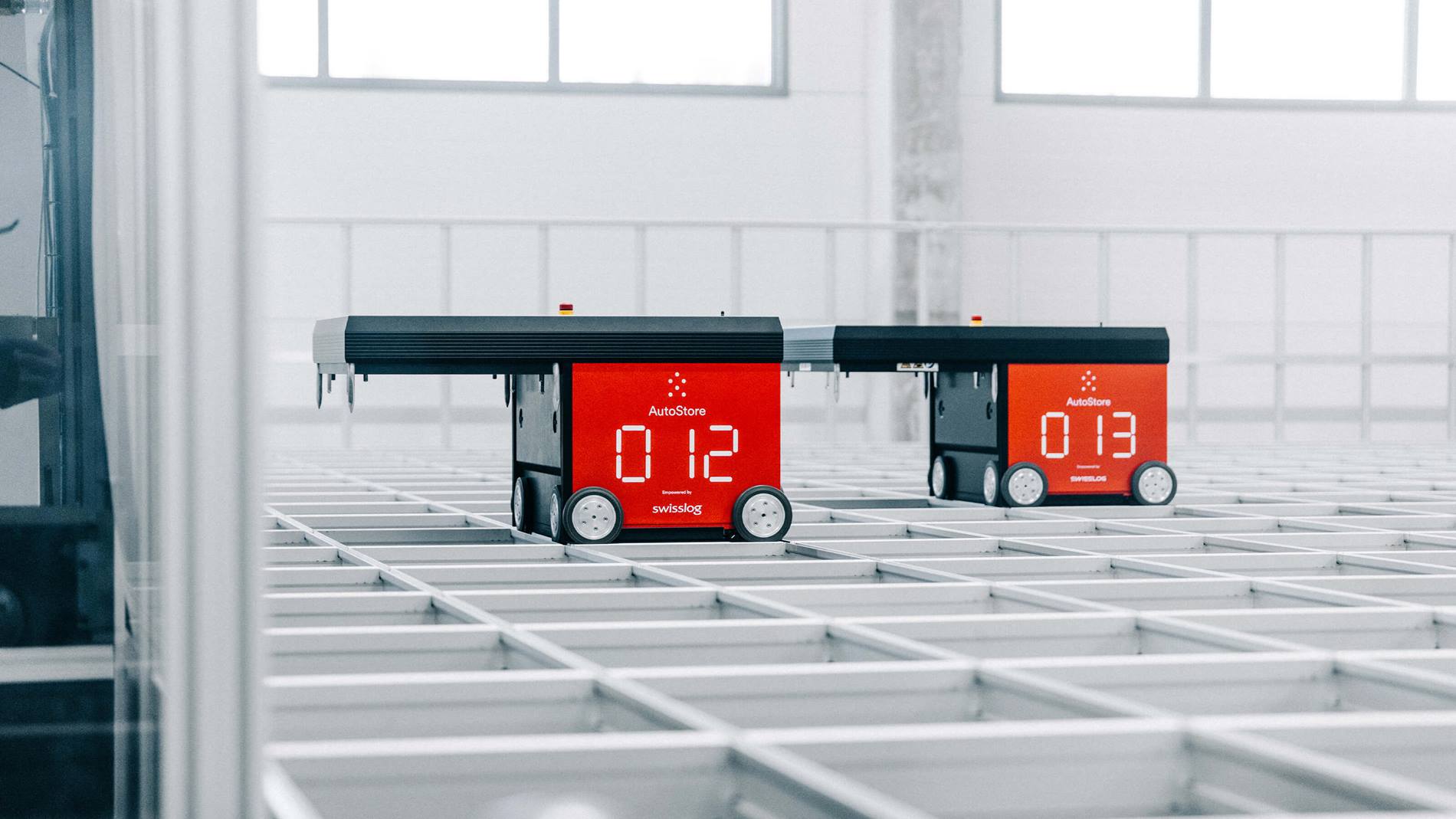 Two red AutoStore robots, operating on a grid system in a warehouse. The robots are part of an automated storage and retrieval system used by Nordic Lights, a Swisslog customer.